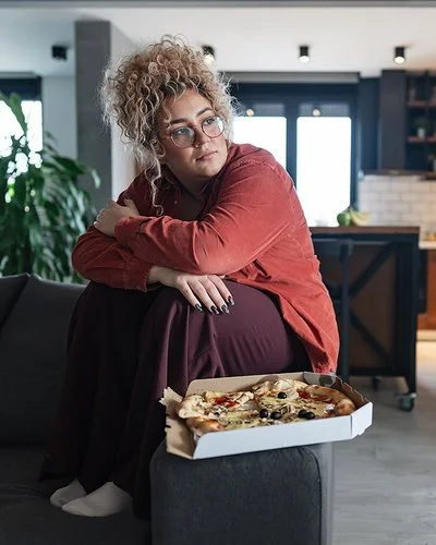 Eine mehrgewichtige Frau sitzt im Wohnzimmer auf dem Sofa, vor ihr steht eine Schachtel mit Pizza.
