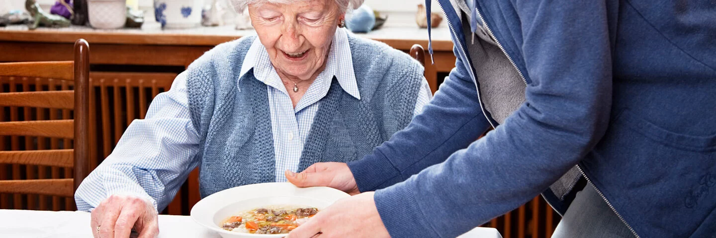 Eine alte Frau sitzt am Tisch und bekommt von einer jüngeren Frau einen Teller mit Essen serviert.