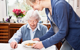 Eine alte Frau sitzt am Tisch und bekommt von einer jüngeren Frau einen Teller mit Essen serviert.