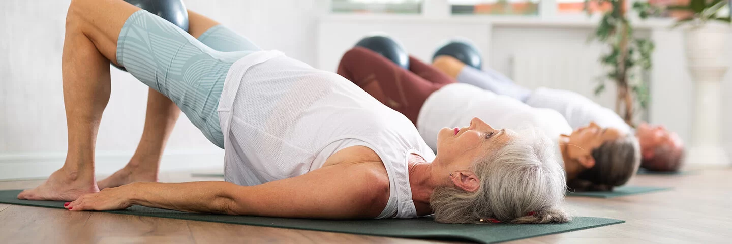Eine Gruppe älterer Frauen turnt auf Yogamatten die Position „die Brücke“.