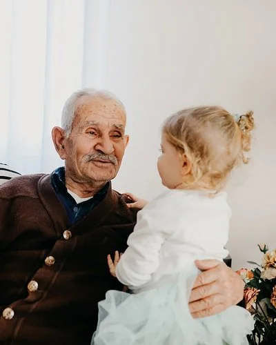 Auf einem Sofa in einem Wohnzimmer sitzen eine junge Frau, ein alter Mann und ein kleines Mädchen. Der alte Mann hat den Arm um das Kind gelegt und lächelt es an. Die Frau sitzt mit einem ebenfalls lächelnden Gesicht daneben.