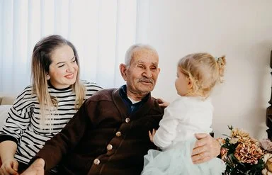 Auf einem Sofa in einem Wohnzimmer sitzen eine junge Frau, ein alter Mann und ein kleines Mädchen. Der alte Mann hat den Arm um das Kind gelegt und lächelt es an. Die Frau sitzt mit einem ebenfalls lächelnden Gesicht daneben.