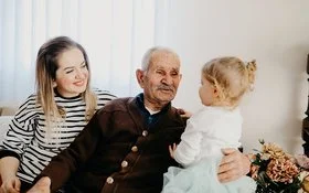 Auf einem Sofa in einem Wohnzimmer sitzen eine junge Frau, ein alter Mann und ein kleines Mädchen. Der alte Mann hat den Arm um das Kind gelegt und lächelt es an. Die Frau sitzt mit einem ebenfalls lächelnden Gesicht daneben.