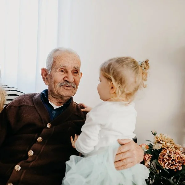 Auf einem Sofa in einem Wohnzimmer sitzen eine junge Frau, ein alter Mann und ein kleines Mädchen. Der alte Mann hat den Arm um das Kind gelegt und lächelt es an. Die Frau sitzt mit einem ebenfalls lächelnden Gesicht daneben.