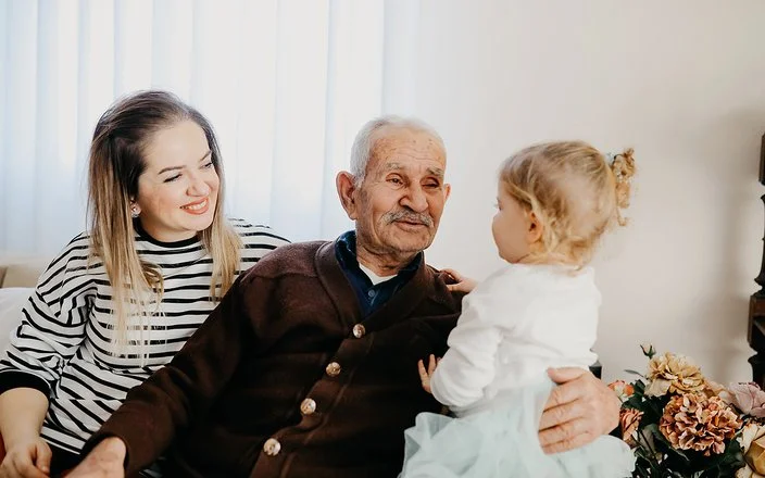 Auf einem Sofa in einem Wohnzimmer sitzen eine junge Frau, ein alter Mann und ein kleines Mädchen. Der alte Mann hat den Arm um das Kind gelegt und lächelt es an. Die Frau sitzt mit einem ebenfalls lächelnden Gesicht daneben.