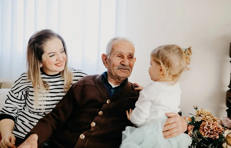 Auf einem Sofa in einem Wohnzimmer sitzen eine junge Frau, ein alter Mann und ein kleines Mädchen. Der alte Mann hat den Arm um das Kind gelegt und lächelt es an. Die Frau sitzt mit einem ebenfalls lächelnden Gesicht daneben.