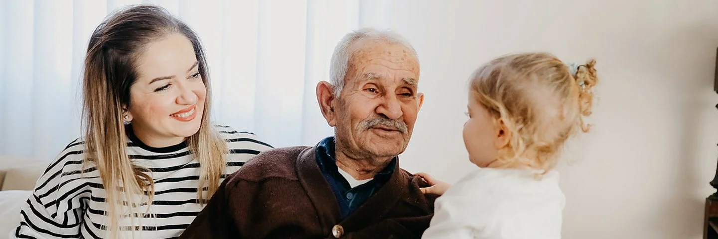 Auf einem Sofa in einem Wohnzimmer sitzen eine junge Frau, ein alter Mann und ein kleines Mädchen. Der alte Mann hat den Arm um das Kind gelegt und lächelt es an. Die Frau sitzt mit einem ebenfalls lächelnden Gesicht daneben.