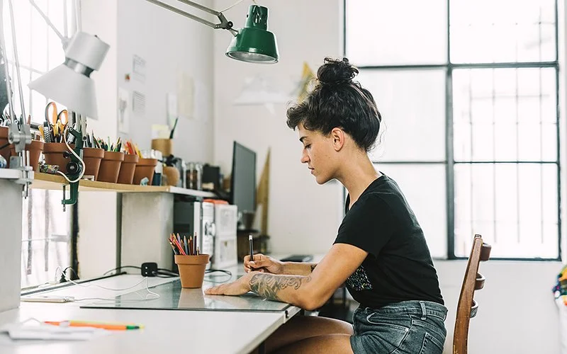 Eine junge Frau sitzt mit kurzen Jeans und in einem schwarzem T-Shirt am Schreibtisch in einem modernen, kreativen Büro oder Ateilier und zeichnet oder schreibt etwas mit einem Stift.