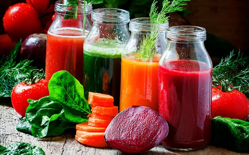 Vier verschiedene Obst- und Gemüsesäfte in rot, grün, orange und dunkelrot, die man bei einer Saftkur zu sich nehmen kann, stehen auf einem Tisch.