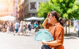 Eine Frau steht im Vordergrund auf einem sonnigen Platz. Sie trägt ein braunes Hemd und hält einen bunt gemusterten Fächer in der rechten Hand. Ihre linke Hand liegt an der Stirn, als würde sie sich vor der Sonne schützen. Im Hintergrund sind mehrere Menschen zu sehen, die an Tischen unter weißen Sonnenschirmen sitzen oder vorbeigehen. Ein grüner Baum spendet etwas Schatten. Die Szene wirkt sommerlich und hell.