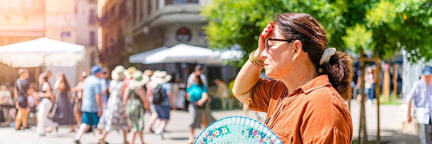 Eine Frau steht im Vordergrund auf einem sonnigen Platz. Sie trägt ein braunes Hemd und hält einen bunt gemusterten Fächer in der rechten Hand. Ihre linke Hand liegt an der Stirn, als würde sie sich vor der Sonne schützen. Im Hintergrund sind mehrere Menschen zu sehen, die an Tischen unter weißen Sonnenschirmen sitzen oder vorbeigehen. Ein grüner Baum spendet etwas Schatten. Die Szene wirkt sommerlich und hell.