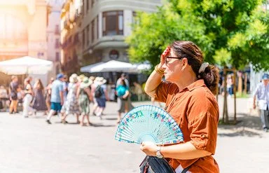 Eine Frau steht im Vordergrund auf einem sonnigen Platz. Sie trägt ein braunes Hemd und hält einen bunt gemusterten Fächer in der rechten Hand. Ihre linke Hand liegt an der Stirn, als würde sie sich vor der Sonne schützen. Im Hintergrund sind mehrere Menschen zu sehen, die an Tischen unter weißen Sonnenschirmen sitzen oder vorbeigehen. Ein grüner Baum spendet etwas Schatten. Die Szene wirkt sommerlich und hell.