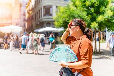Eine Frau steht im Vordergrund auf einem sonnigen Platz. Sie trägt ein braunes Hemd und hält einen bunt gemusterten Fächer in der rechten Hand. Ihre linke Hand liegt an der Stirn, als würde sie sich vor der Sonne schützen. Im Hintergrund sind mehrere Menschen zu sehen, die an Tischen unter weißen Sonnenschirmen sitzen oder vorbeigehen. Ein grüner Baum spendet etwas Schatten. Die Szene wirkt sommerlich und hell.