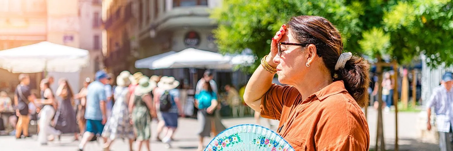 Eine Frau steht im Vordergrund auf einem sonnigen Platz. Sie trägt ein braunes Hemd und hält einen bunt gemusterten Fächer in der rechten Hand. Ihre linke Hand liegt an der Stirn, als würde sie sich vor der Sonne schützen. Im Hintergrund sind mehrere Menschen zu sehen, die an Tischen unter weißen Sonnenschirmen sitzen oder vorbeigehen. Ein grüner Baum spendet etwas Schatten. Die Szene wirkt sommerlich und hell.