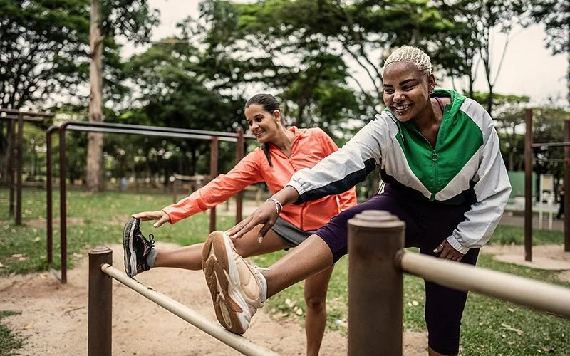 Zwei junge Frauen in sportlicher Kleidung dehnen ihre Beine an einer Stange in einem Park.