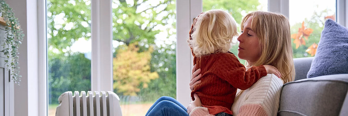 Mutter sitzt mit einem Kleinkind auf dem Schoss auf dem Teppich und lehnt an der Couch.