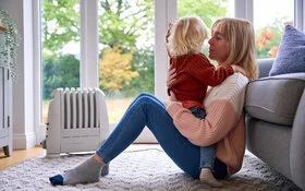 Mutter sitzt mit einem Kleinkind auf dem Schoss auf dem Teppich und lehnt an der Couch.