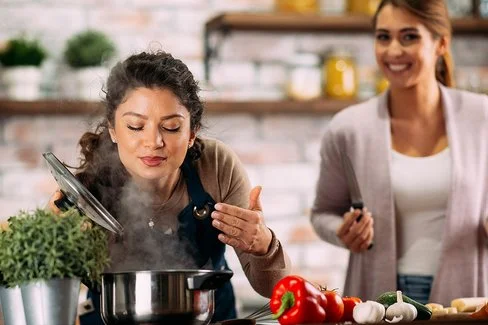 Zwei Frauen stehen in einer Küche, eine riecht an einem dampfenden Topf während die andere neben ihr steht und Zutaten vorbereitet