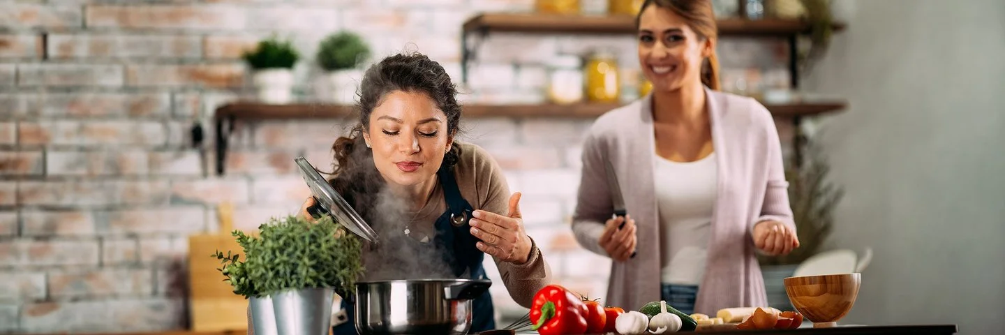Zwei Frauen stehen in einer Küche, eine riecht an einem dampfenden Topf während die andere neben ihr steht und Zutaten vorbereitet