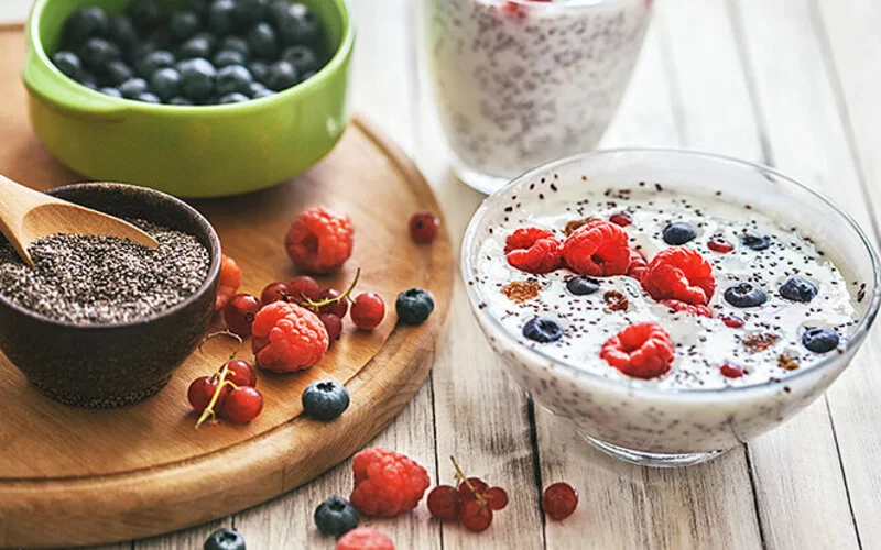 Auf einem Tisch liegen Chiasamen, Blaubeeren, Himbeeren und ein frisch zubereiteter Joghurt