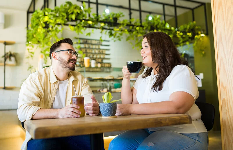 Ein Mann und eine Frau, die beide von Übergewicht betroffen sind, sitzen in einem Café. Die Frau trinkt einen Kaffee, der Mann einen Eistee.