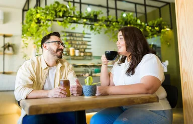 Ein Mann und eine Frau, die beide von Übergewicht betroffen sind, sitzen in einem Café. Die Frau trinkt einen Kaffee, der Mann einen Eistee.