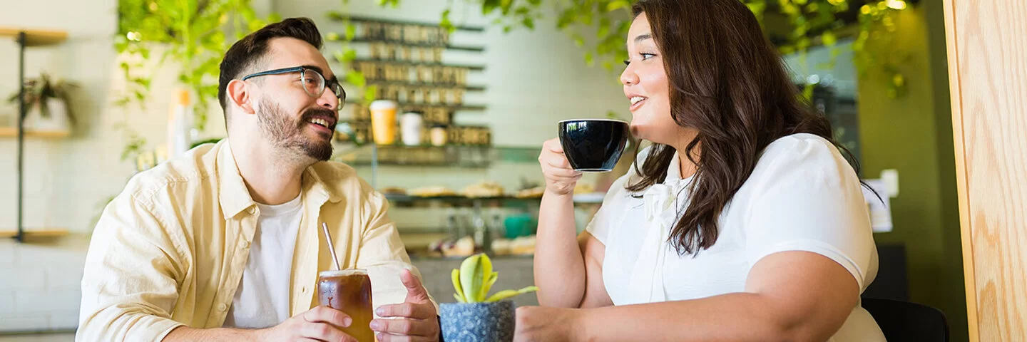 Ein Mann und eine Frau, die beide von Übergewicht betroffen sind, sitzen in einem Café. Die Frau trinkt einen Kaffee, der Mann einen Eistee.