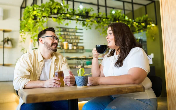 Ein Mann und eine Frau, die beide von Übergewicht betroffen sind, sitzen in einem Café. Die Frau trinkt einen Kaffee, der Mann einen Eistee.