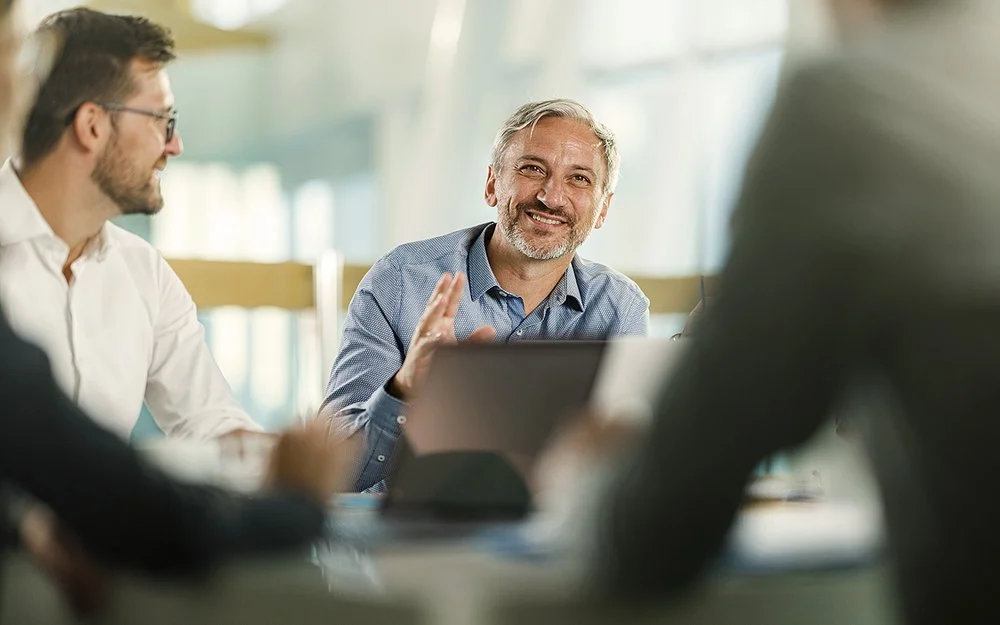 An einem Tisch in einem Büro sitzen vier Personen. Ein Mann ist im Profil, ein anderer frontal aufgenommen. Beide lächeln. Zwei Personen sind nur ausschnittsweise und verschwommen erfasst.