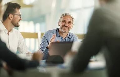 An einem Tisch in einem Büro sitzen vier Personen. Ein Mann ist im Profil, ein anderer frontal aufgenommen. Beide lächeln. Zwei Personen sind nur ausschnittsweise und verschwommen erfasst.