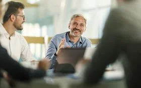 An einem Tisch in einem Büro sitzen vier Personen. Ein Mann ist im Profil, ein anderer frontal aufgenommen. Beide lächeln. Zwei Personen sind nur ausschnittsweise und verschwommen erfasst.