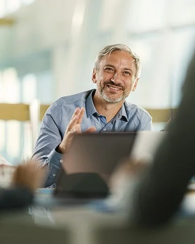 An einem Tisch in einem Büro sitzen vier Personen. Ein Mann ist im Profil, ein anderer frontal aufgenommen. Beide lächeln. Zwei Personen sind nur ausschnittsweise und verschwommen erfasst.