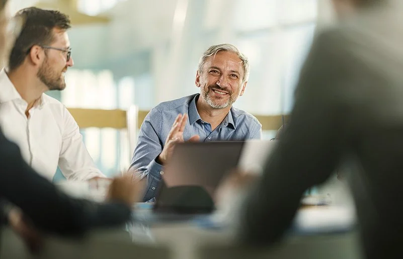 An einem Tisch in einem Büro sitzen vier Personen. Ein Mann ist im Profil, ein anderer frontal aufgenommen. Beide lächeln. Zwei Personen sind nur ausschnittsweise und verschwommen erfasst.