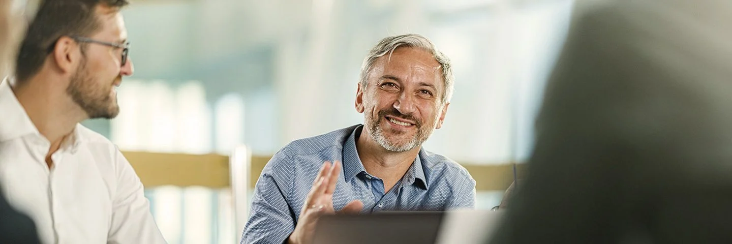 An einem Tisch in einem Büro sitzen vier Personen. Ein Mann ist im Profil, ein anderer frontal aufgenommen. Beide lächeln. Zwei Personen sind nur ausschnittsweise und verschwommen erfasst.