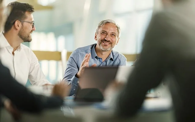 An einem Tisch in einem Büro sitzen vier Personen. Ein Mann ist im Profil, ein anderer frontal aufgenommen. Beide lächeln. Zwei Personen sind nur ausschnittsweise und verschwommen erfasst.