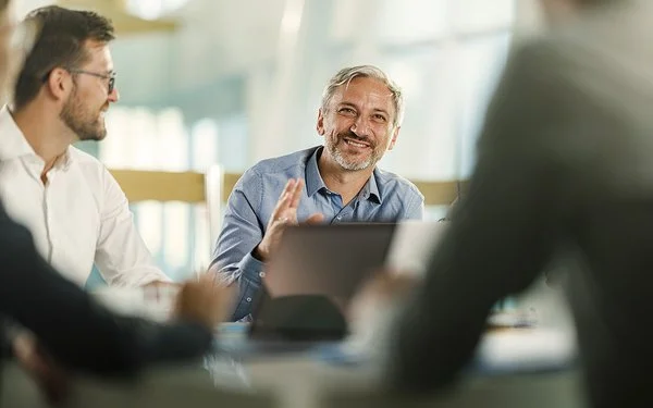 An einem Tisch in einem Büro sitzen vier Personen. Ein Mann ist im Profil, ein anderer frontal aufgenommen. Beide lächeln. Zwei Personen sind nur ausschnittsweise und verschwommen erfasst.