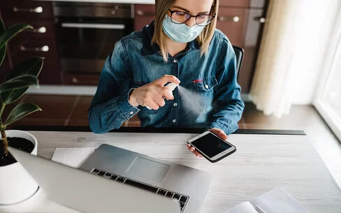Eine Frau mit Maske sitzt vor ihrem Laptop am Schreibtisch und desinfiziert mit einem Spray ihr Mobiltelefon.