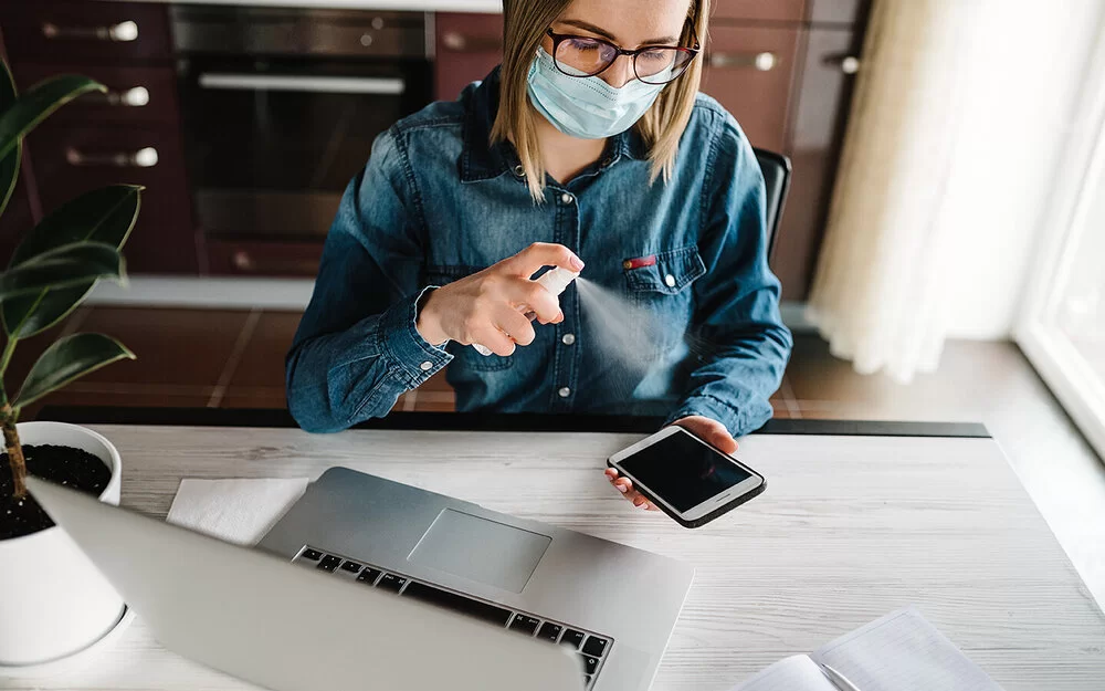 Eine Frau mit Maske sitzt vor ihrem Laptop am Schreibtisch und desinfiziert mit einem Spray ihr Mobiltelefon.