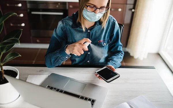 Eine Frau mit Maske sitzt vor ihrem Laptop am Schreibtisch und desinfiziert mit einem Spray ihr Mobiltelefon.
