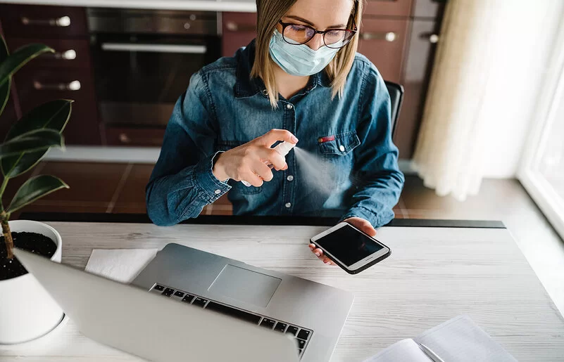 Eine Frau mit Maske sitzt vor ihrem Laptop am Schreibtisch und desinfiziert mit einem Spray ihr Mobiltelefon.