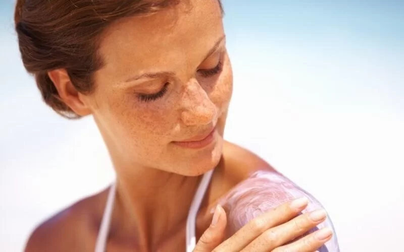 Eine junge Frau mit Sommersprossen cremt sich ihre Schulter mit Sonnenmilch ein.
