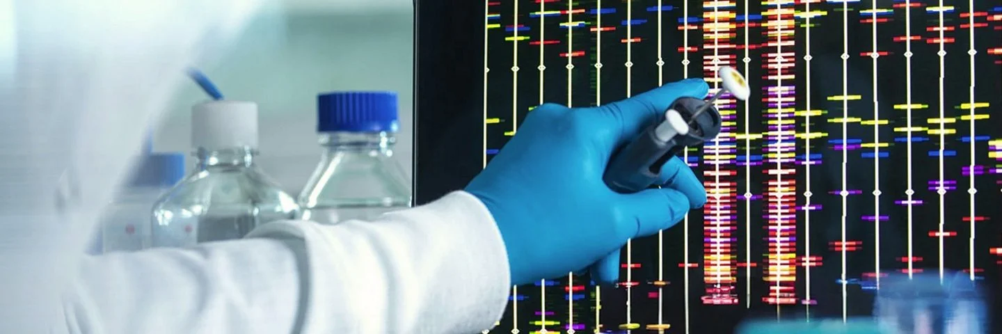 In einem Labor stehen Probebehälter vor einem Monitor, auf dem farbige Balken an senkrechten Linien angeordnet sind. Eine Hand im Kunststoffhandschuh zeigt auf den Monitor.