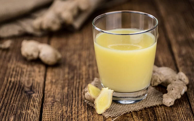 Ein Glas Ingwer-Shot wird neben Ingwerknollen auf einem Holztisch präsentiert.