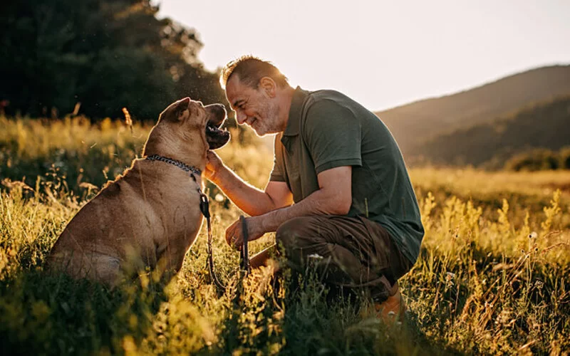 Ein älterer Mann hockt neben seinem Hund auf einem Feld.