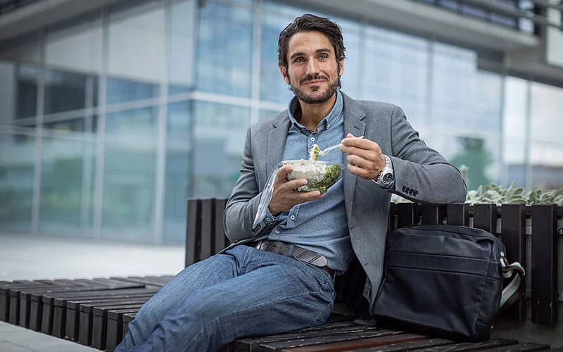 Ein junger Mann macht Mittagspause auf einer Bank vor dem Bürogebäude.