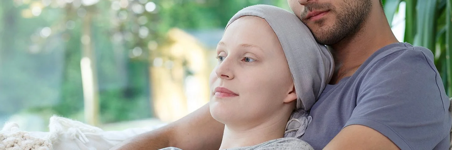 Ein Mann hält seine Frau, die eine Krebsbehandlung hatte und ihren Kopf mit einem Tuch bedeckt hat, liebevoll in den Armen.