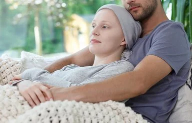 Ein Mann hält seine Frau, die eine Krebsbehandlung hatte und ihren Kopf mit einem Tuch bedeckt hat, liebevoll in den Armen.