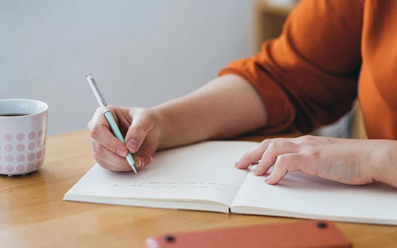 Eine Frau sitzt am Küchentisch und schreibt Notizen zur Stressbewältigung in ein Notizheft.