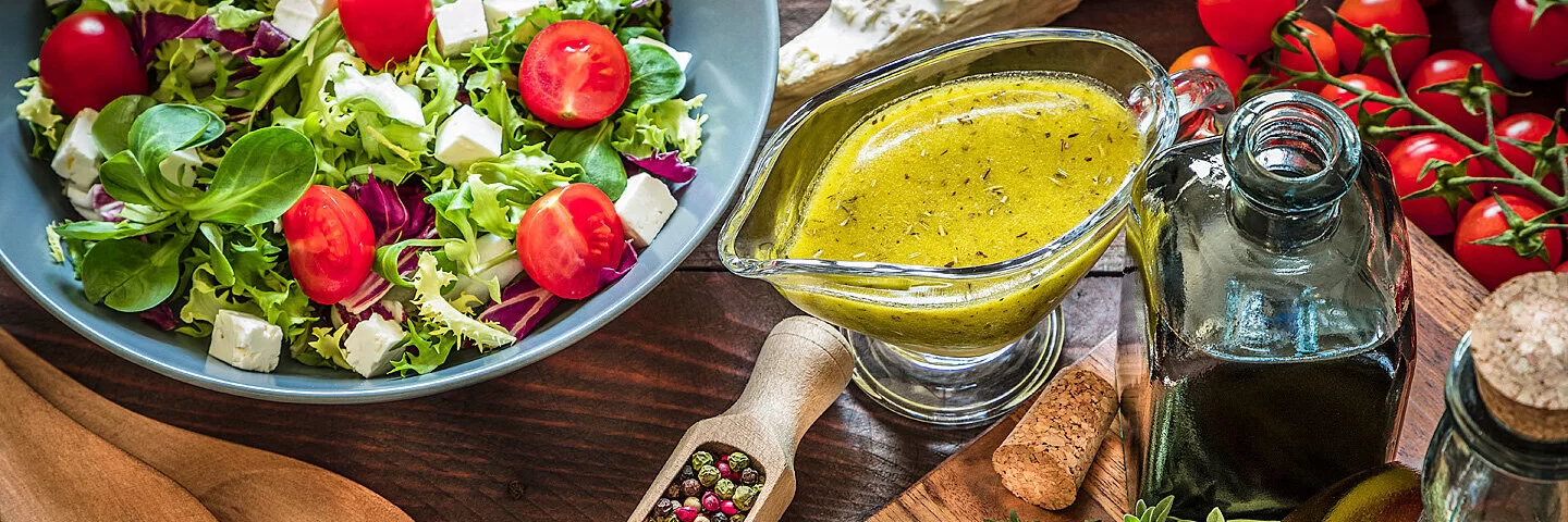 Eine große Schüssel mit gemischten Salat mitsamt einer frisch angemischten Vinaigrette und einer Essigflasche sowie Öl, Kräutern und Gewürzen.