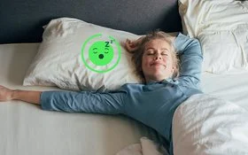 Eine blonde Frau liegt auf dem Rücken in ihrem Bett, die Augen geschlossen und streckt ihren Arm aus. 
